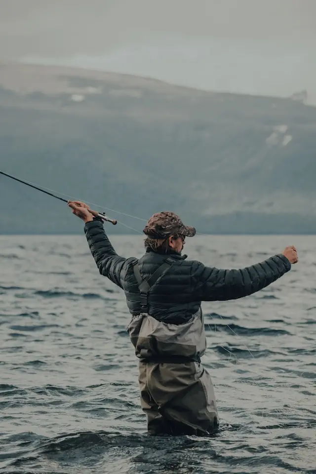 Lystfisker i waders kaster fluestangen ud i naturskønne omgivelser, som symbol på ro og fordybelse ved vandet.