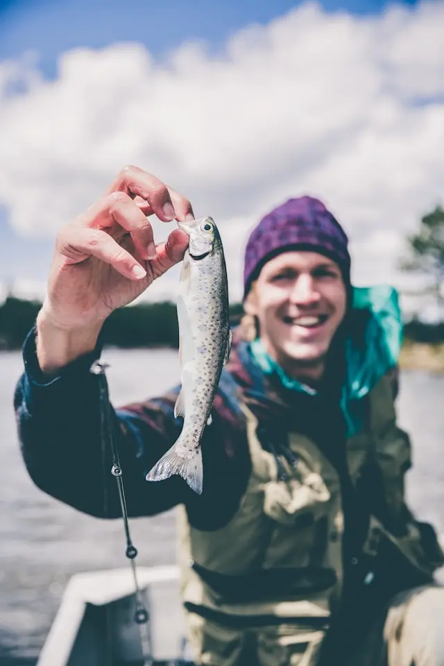 En smilende lystfisker i en lilla hue holder stolt en lille fisk op, som et symbol på fiskeglæde og vores mission.